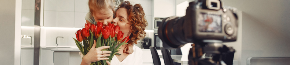 Câmera DSLR em primeiro plano desfocado registrando um ensaio de Dia das Mães afetivo na cozinha, mostrando mãe e filha abraçadas segurando um buquê de tulipas vermelhas.