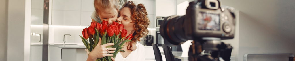 Câmera DSLR em primeiro plano desfocado registrando um ensaio de Dia das Mães afetivo na cozinha, mostrando mãe e filha abraçadas segurando um buquê de tulipas vermelhas.