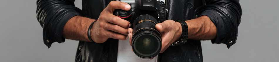 Close nas mãos de um fotógrafo segurando uma câmera DSLR preta com lente profissional, vestindo jaqueta de couro escura.