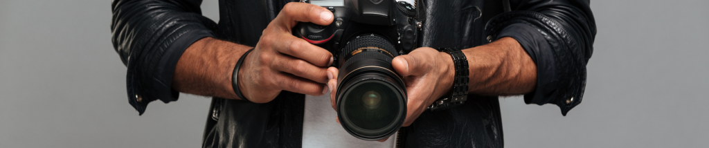 Close nas mãos de um fotógrafo segurando uma câmera DSLR preta com lente profissional, vestindo jaqueta de couro escura.